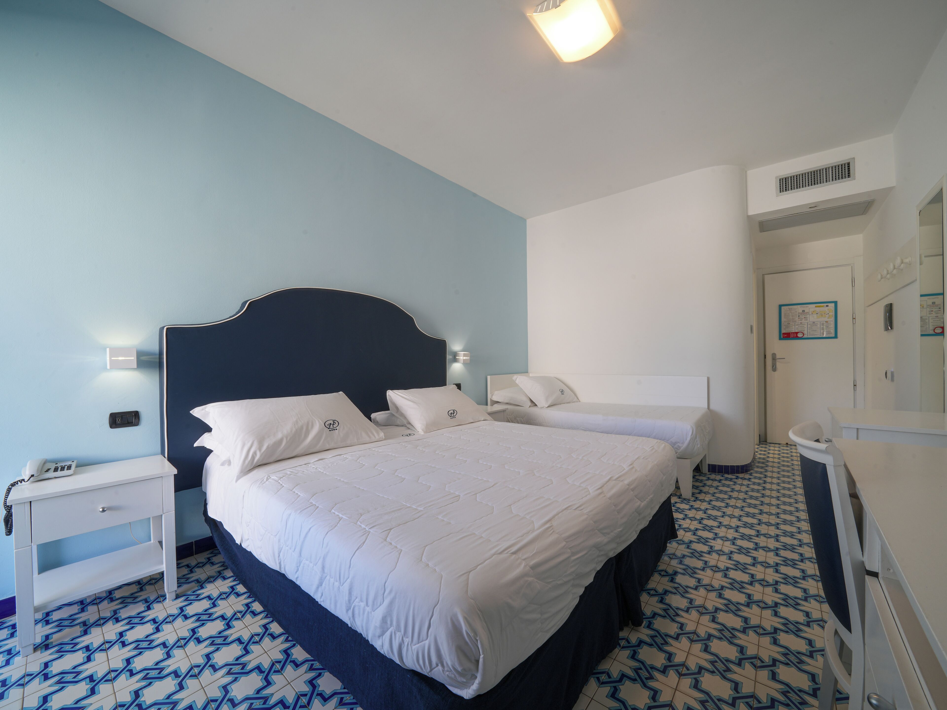 A hotel room with a large bed, two bedside tables, a nightstand, a bathroom with a mirror, and patterned floor tiles.