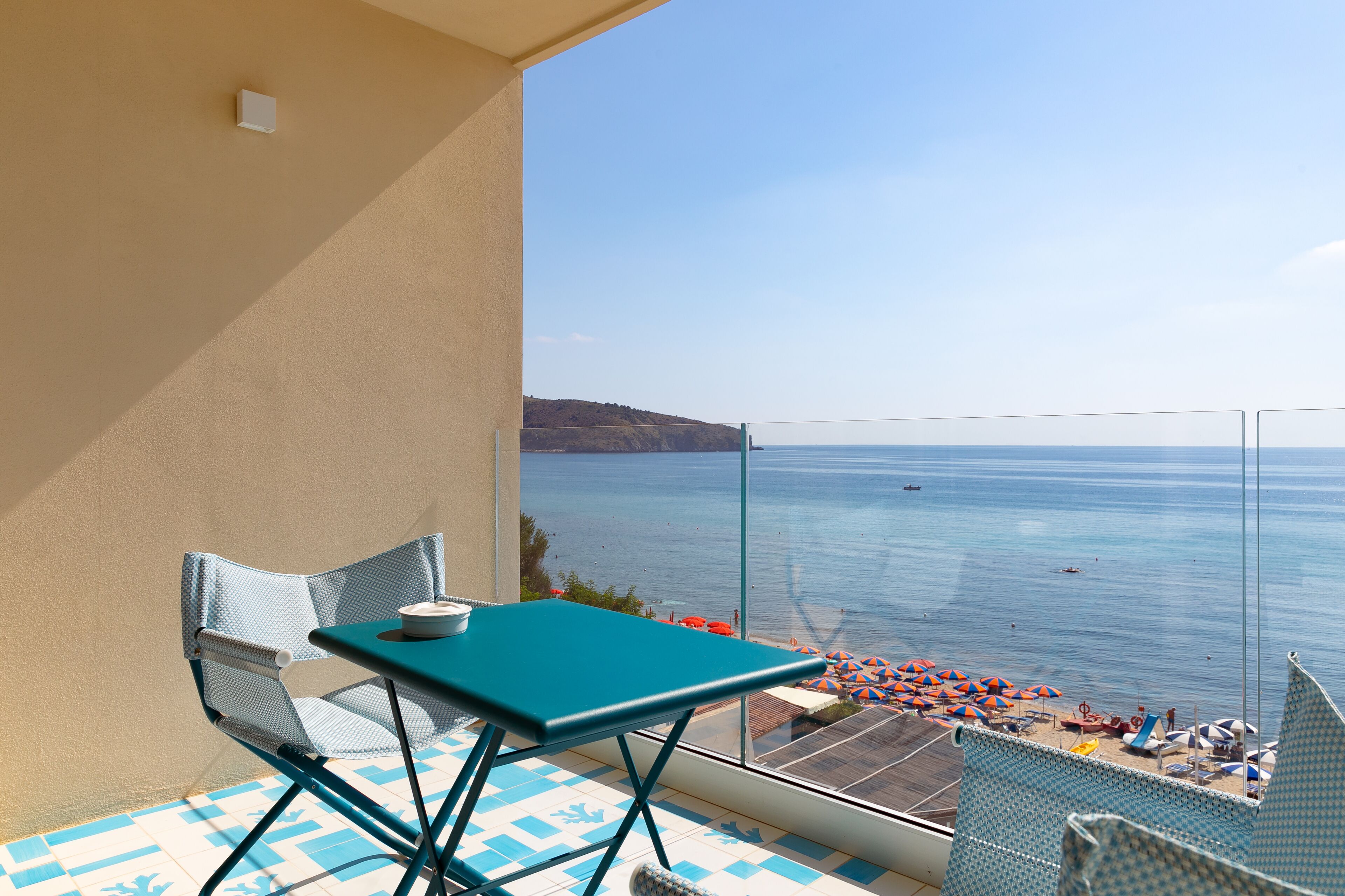 Un balcon avec une table et des chaises, donnant sur une plage parsemée de parasols et offrant une vue dégagée sur la mer.