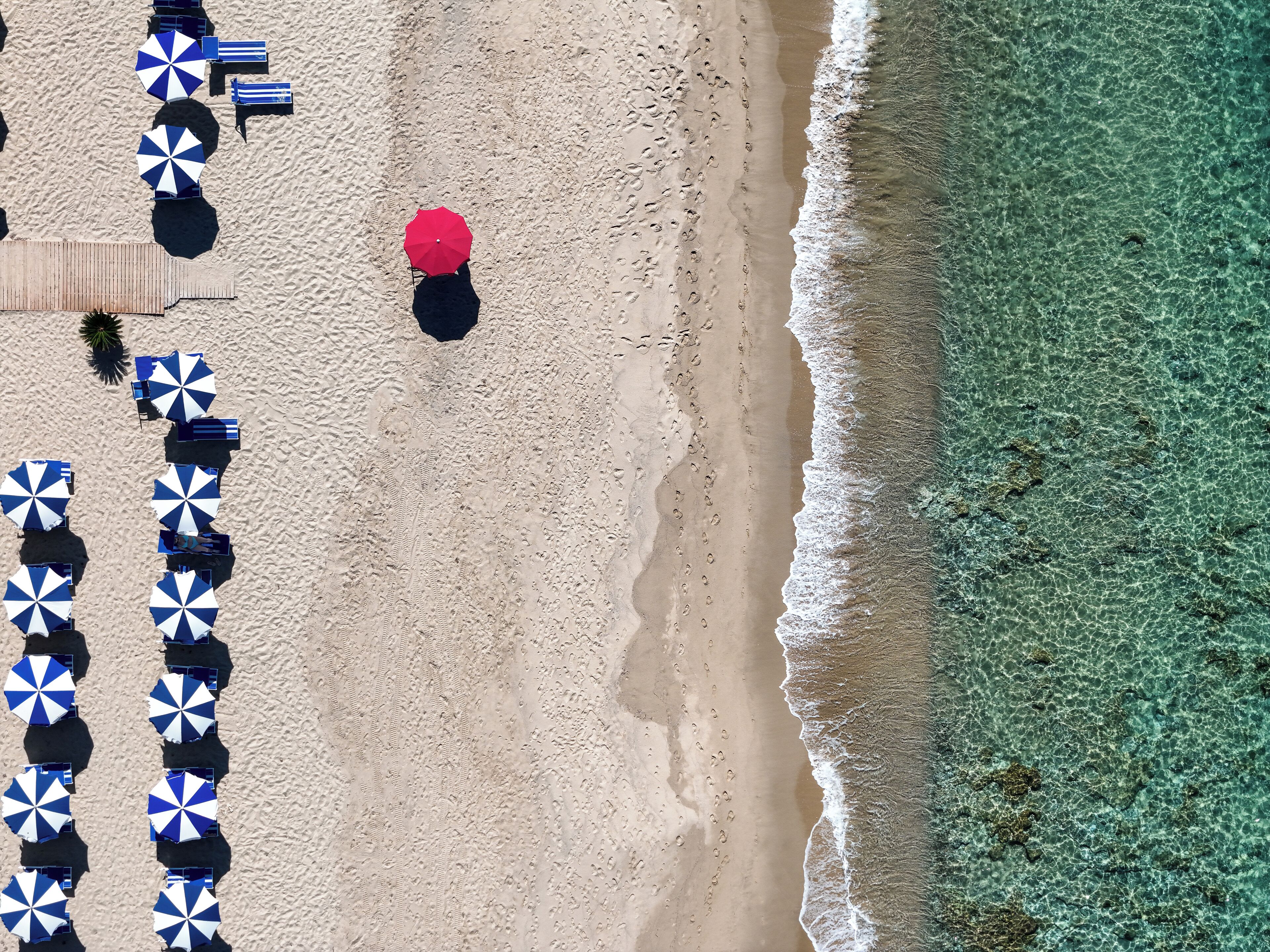 An aerial view of a beach with rows of beach umbrellas and lounge chairs, a wooden walkway, and clear turquoise water.