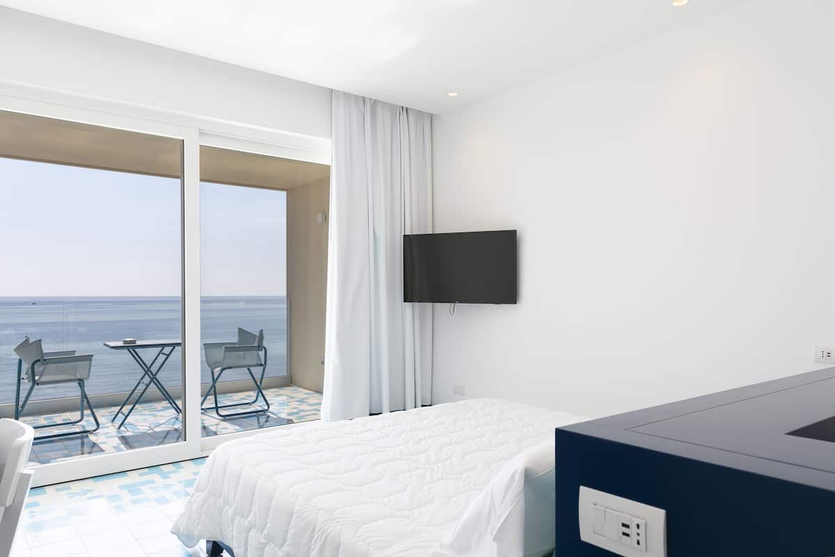 Une chambre d’hôtel avec un lit, une télévision, un balcon avec une table et des chaises, et une vue sur la mer.