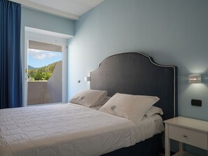 A bedroom with a bed, a nightstand, a wall-mounted light, and a view of greenery through a door. - Hotel Saline Palinuro (Centola)