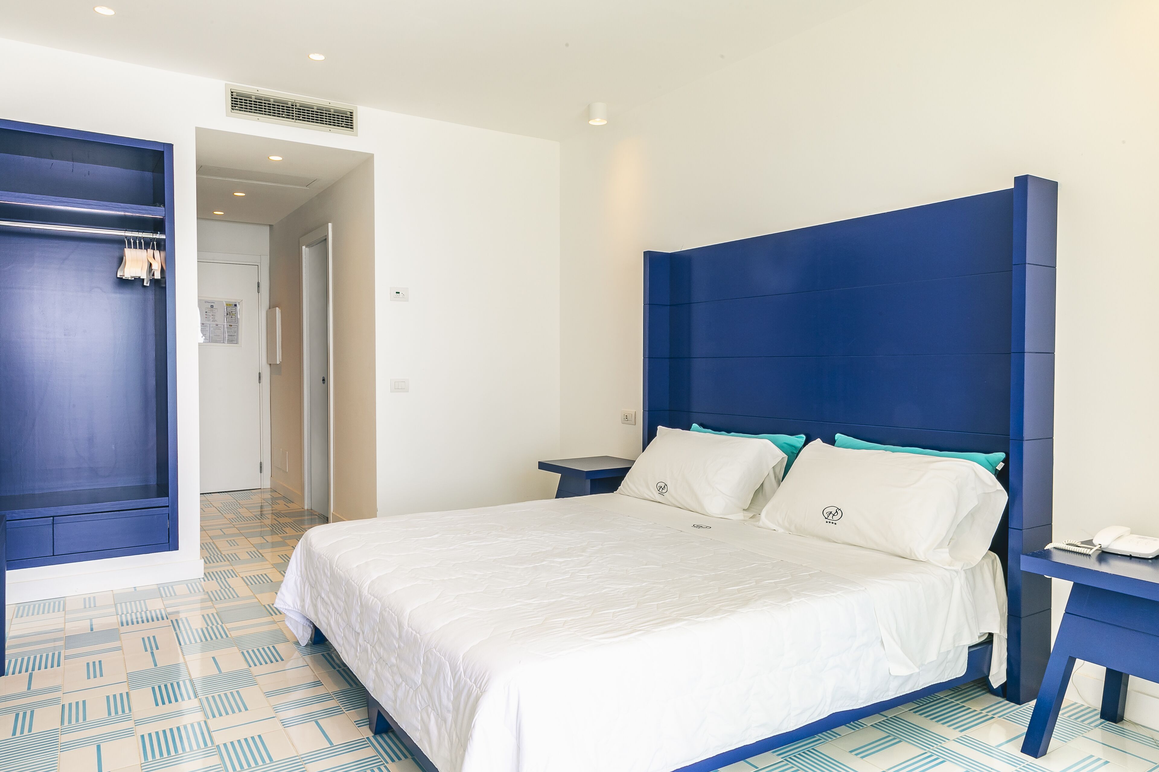 A modern hotel room with a large bed, a blue headboard, and a wardrobe.