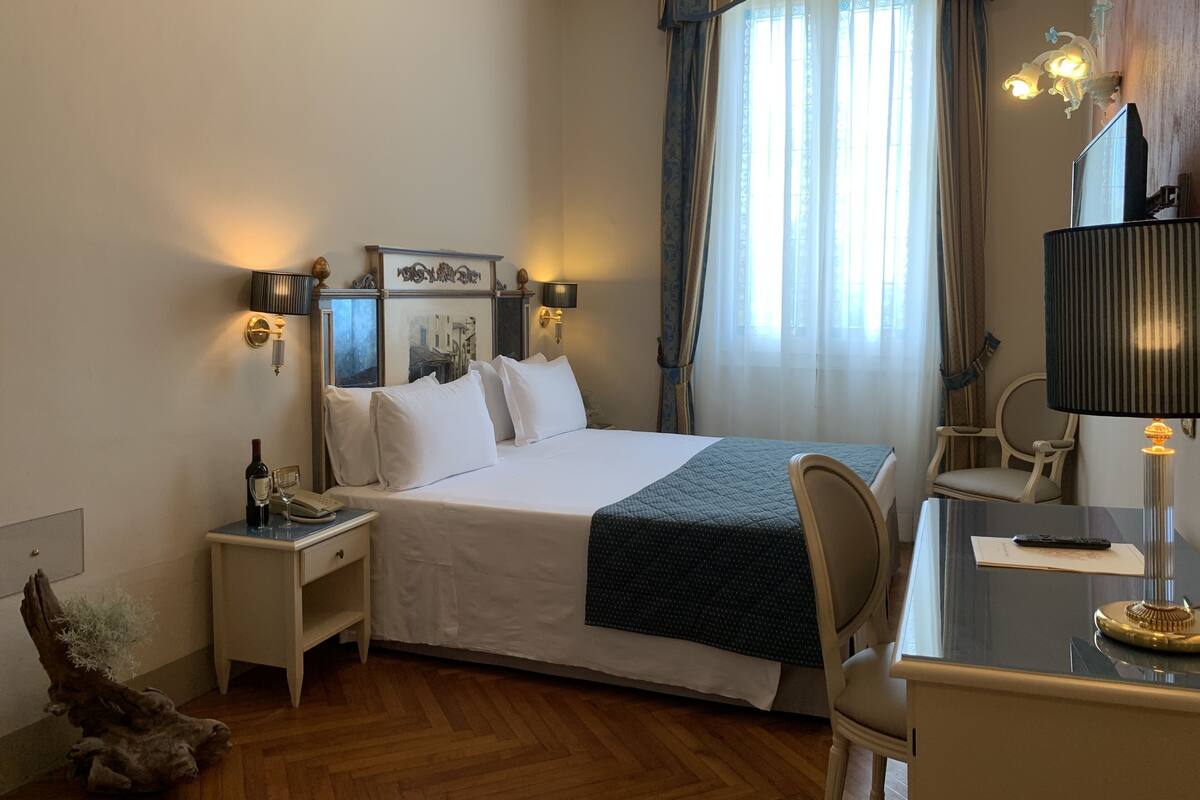 Una habitación de hotel con cama, mesita de noche, lámpara, silla y un gran ventanal con cortinas.