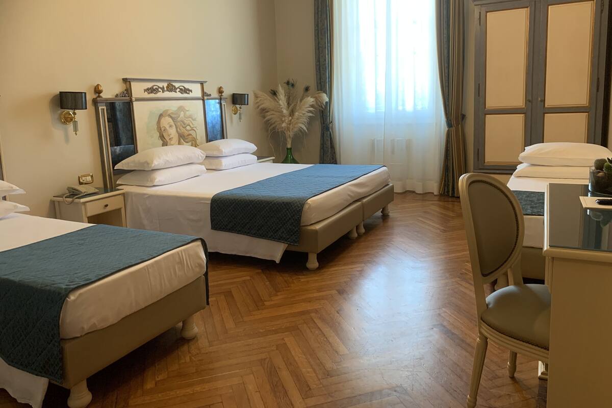 Habitación de hotel con dos camas, un escritorio, una silla, un espejo, un florero con plumas de pampa y una ventana con cortinas.