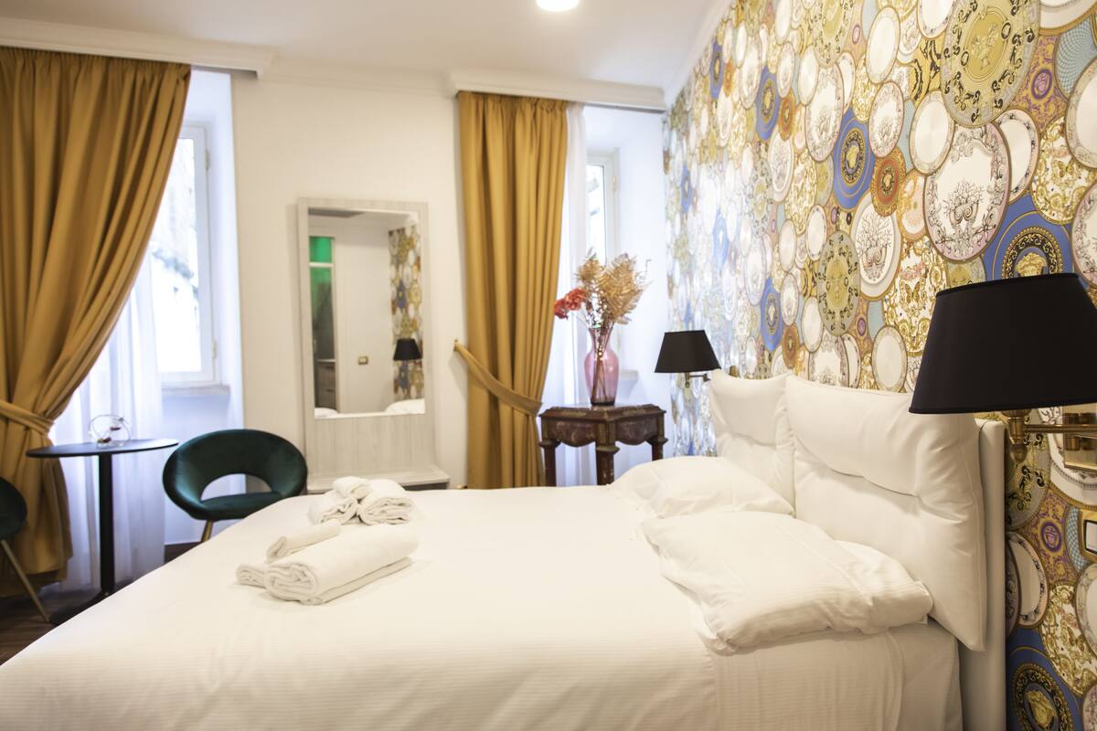 A hotel room with a neatly made bed, a small table with a chair, a mirror, and a wall with decorative plates.