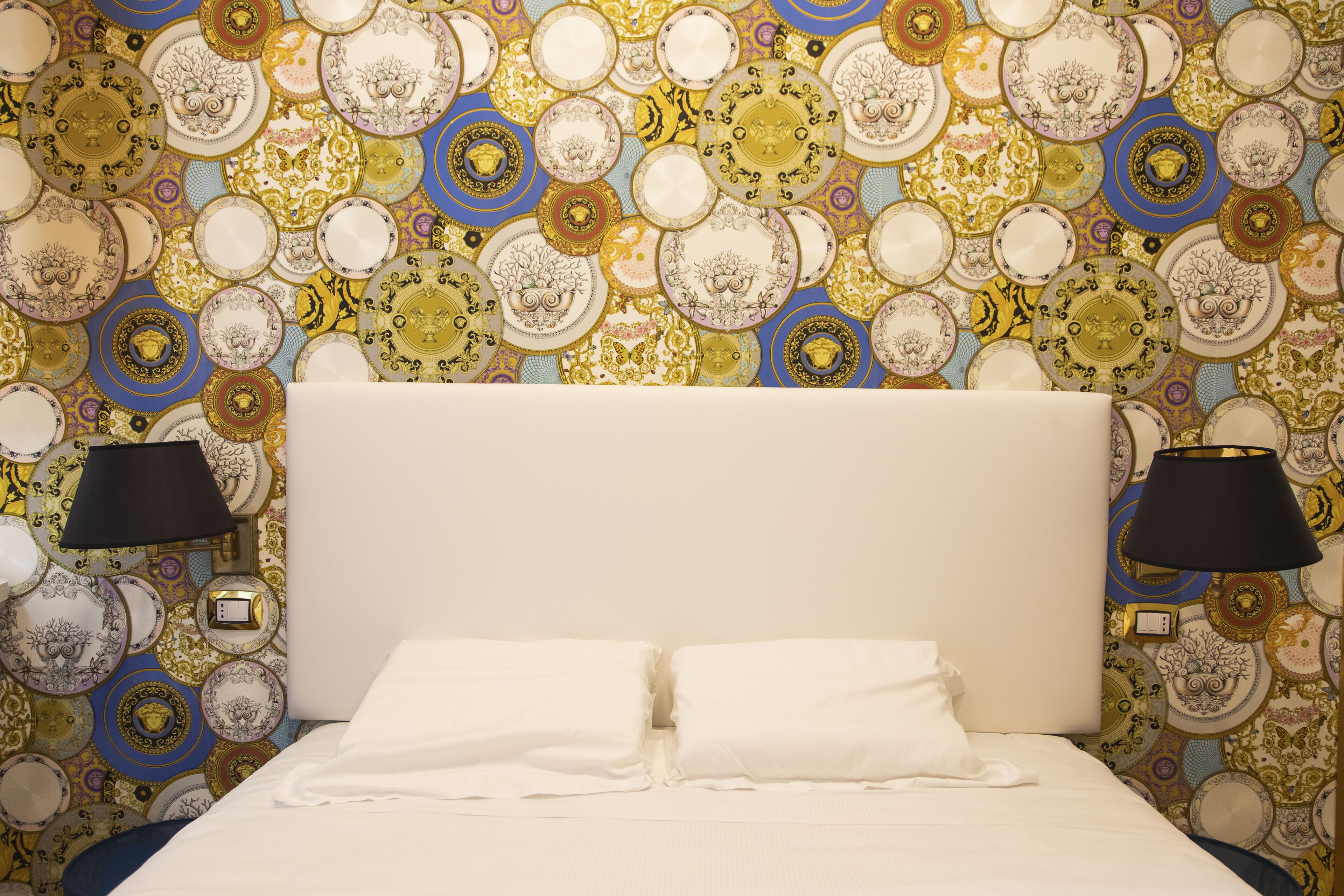 Ein Hotelzimmer mit gemustertem Wandpapier, einem weißen Bett mit zwei Kissen und zwei Wandlampen.
