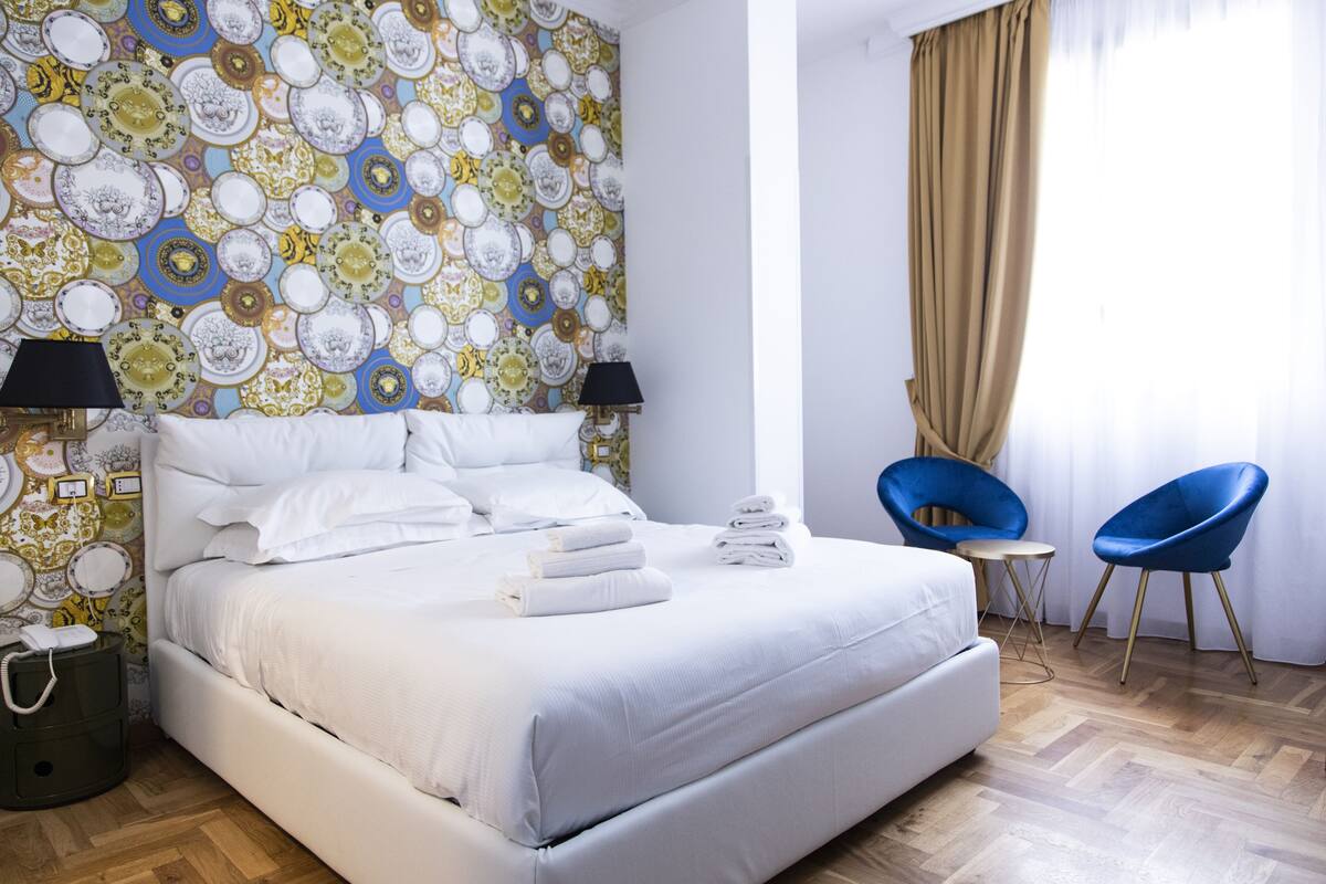 A hotel room with a large bed, two bedside lamps, a blue chair, and a patterned wallpaper.