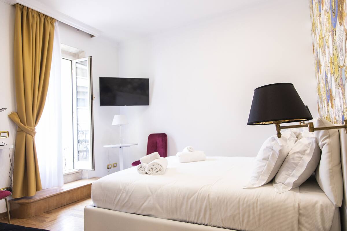 A hotel room with a bed, a television mounted on the wall, a window with curtains, and a lamp.
