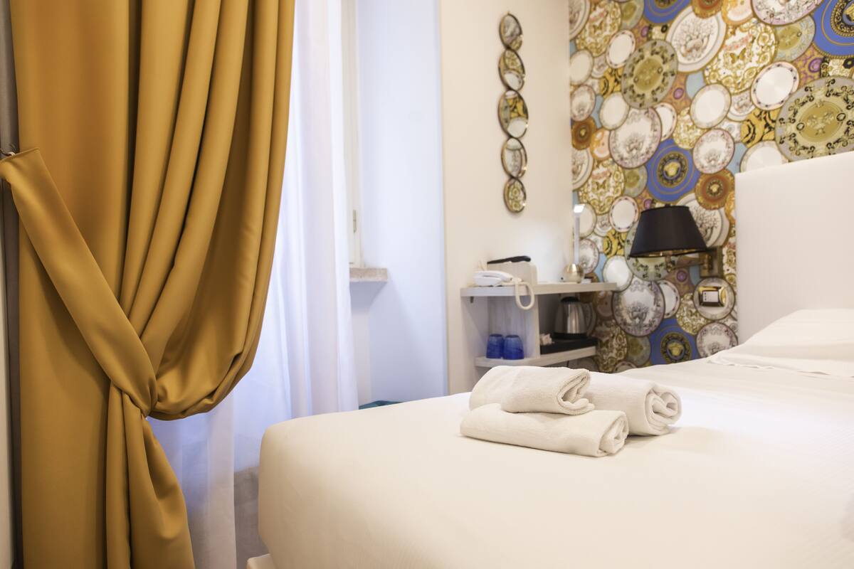 A hotel room with a bed, a bedside table with a lamp, a wall with decorative plates, and a curtain.