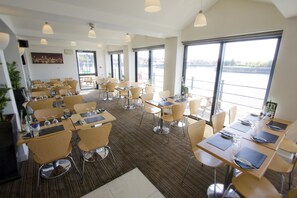 A dining area with multiple tables and chairs, large windows with a view of a body of water, and a modern interior design.