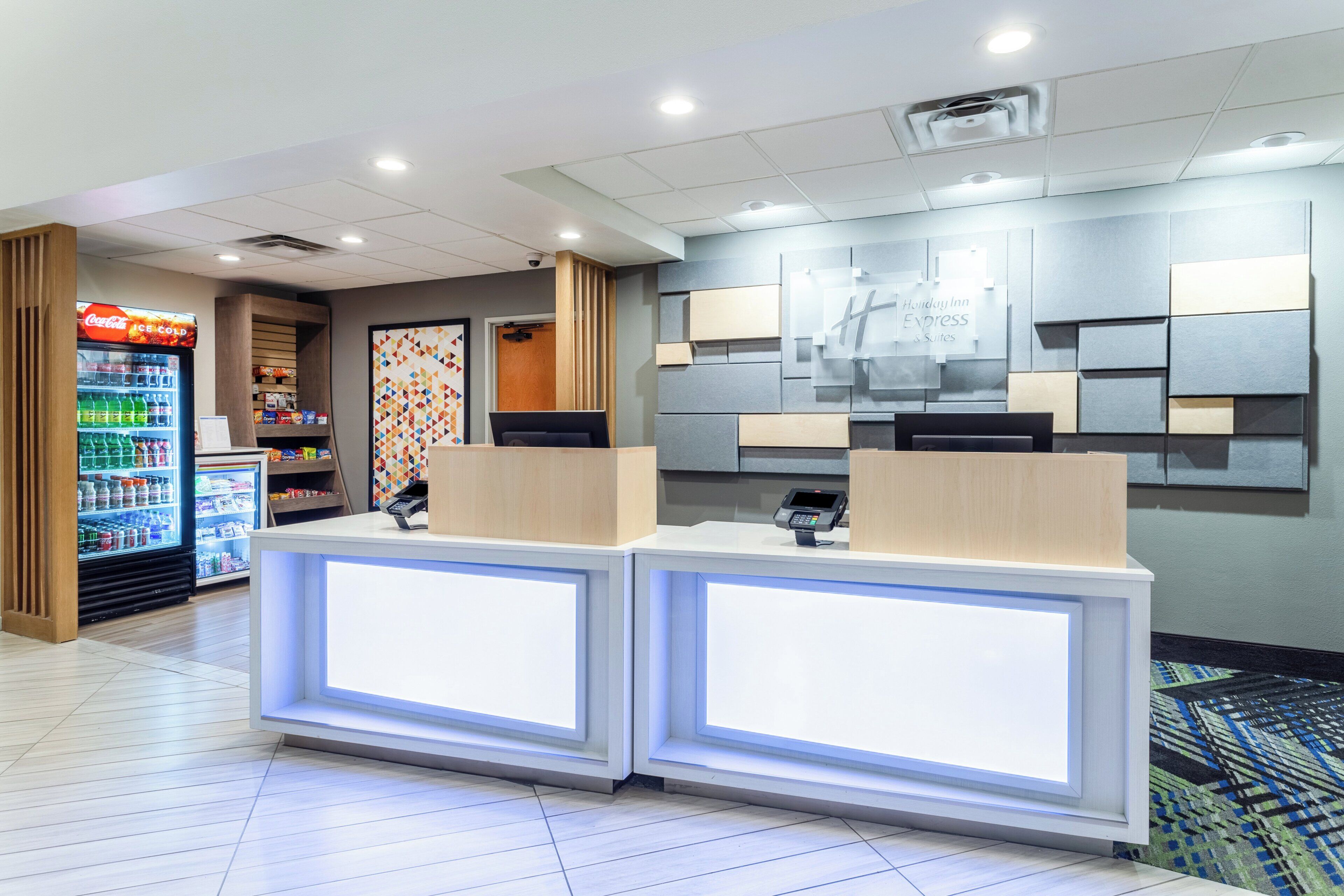 A hotel reception area with a Coca-Cola refrigerator, a digital kiosk, and a wall-mounted sign for Holiday Inn Express & Suites.