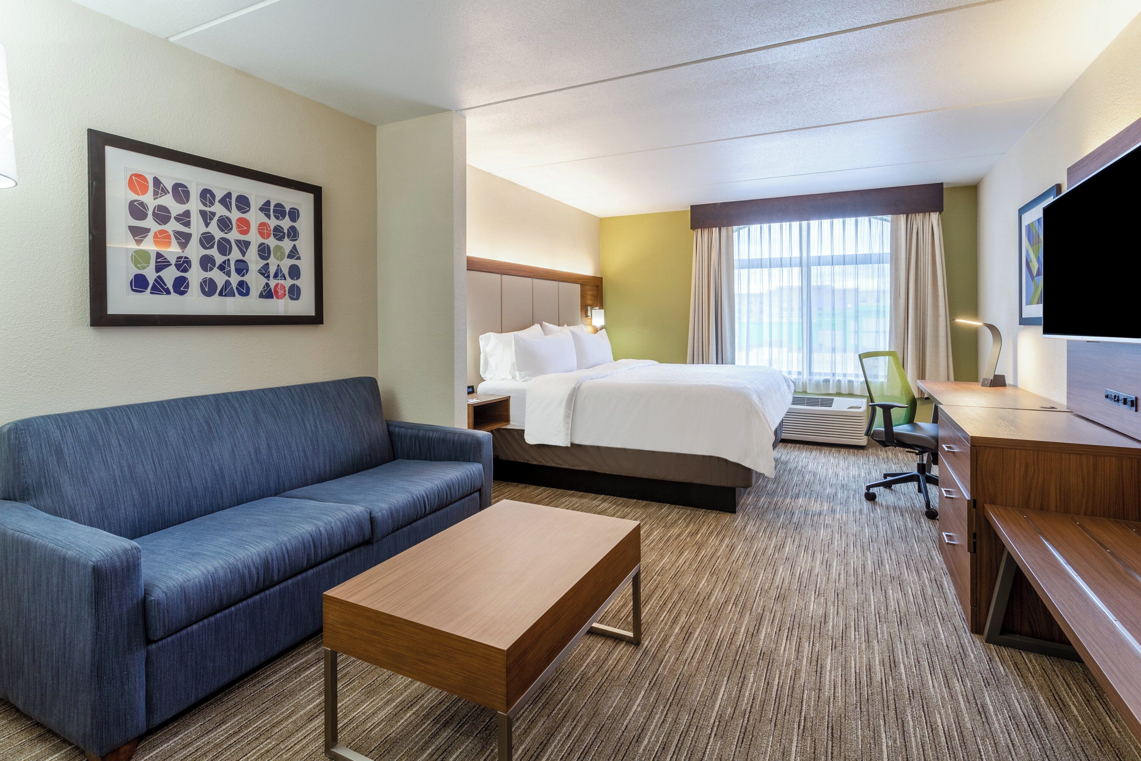 A hotel room with a bed, a sofa, a desk, a TV, and a framed picture on the wall.