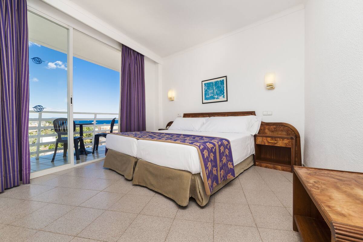 Habitación de hotel con una cama, una mesita de noche de madera, un escritorio y una silla. Hay un balcón con vista al mar.