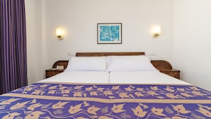 Habitación de hotel con cama, dos mesitas de noche, un atril y un cuadro enmarcado en la pared, además de una cortina púrpura.