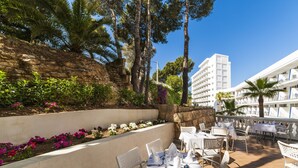 Un área de comedor al aire libre con mesas y sillas blancas, rodeada por un muro de piedra y abundante vegetación.