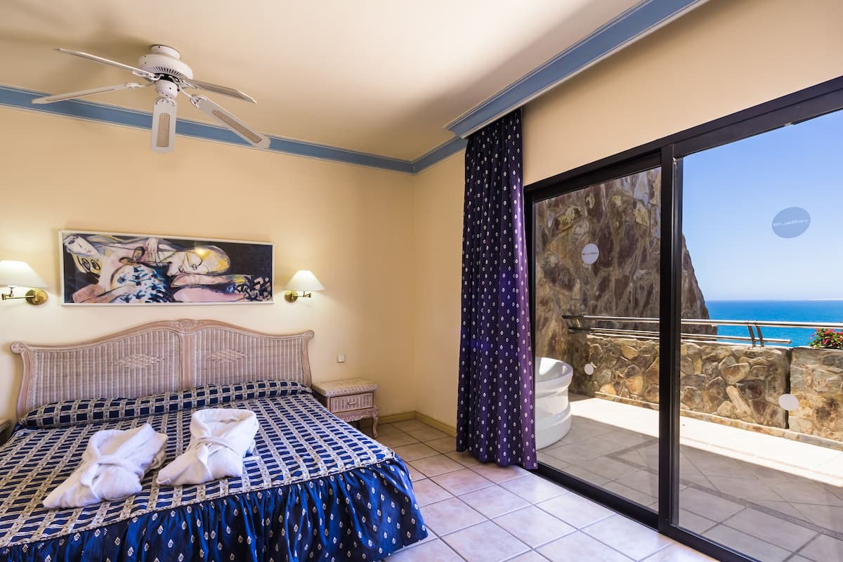 A hotel room with a bed, a ceiling fan, a painting, a nightstand, a bathroom with a bathtub, and a balcony with a view of the sea.