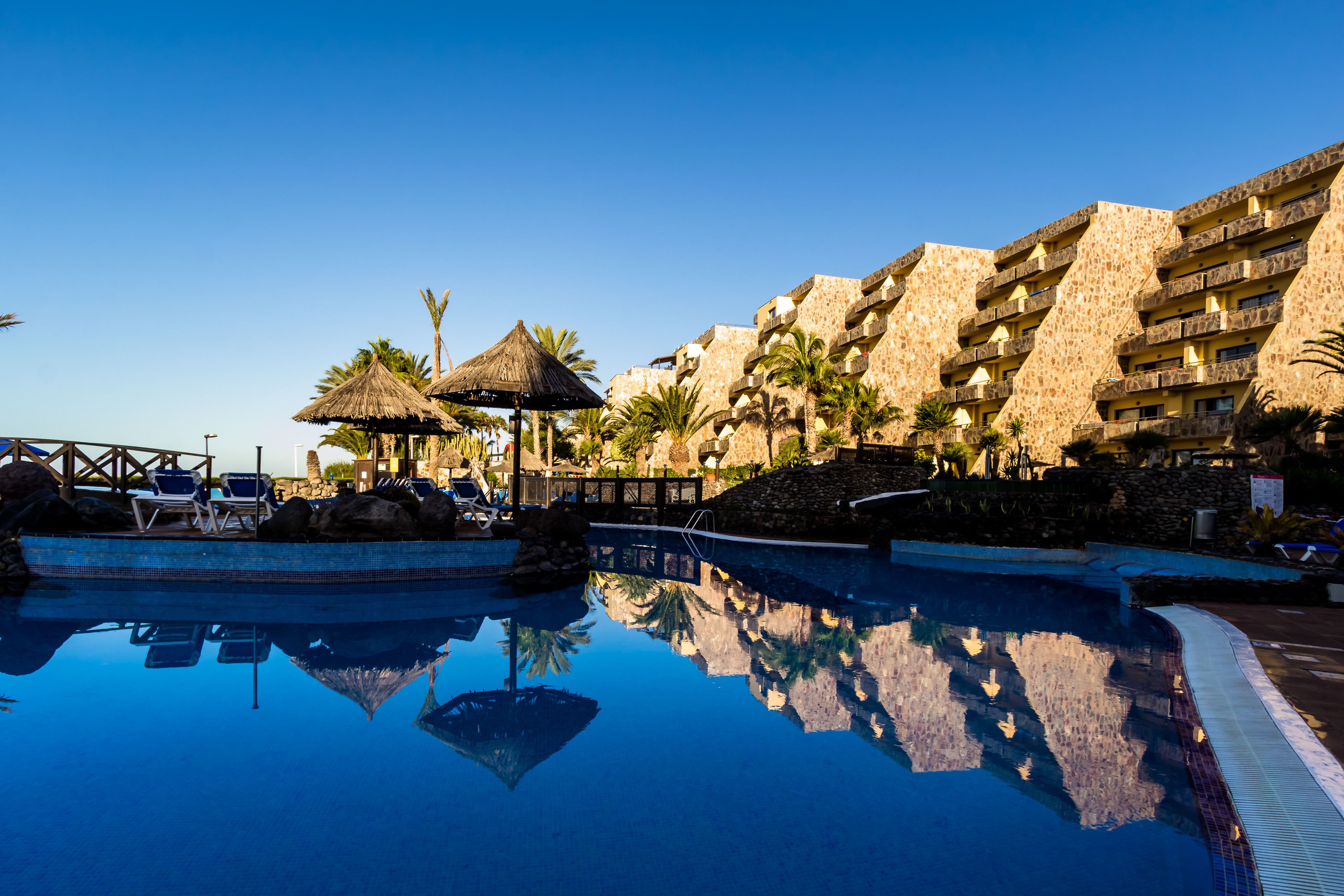 A swimming pool with lounge chairs and a thatched umbrella, surrounded by palm trees and a multi-story building with balconies.