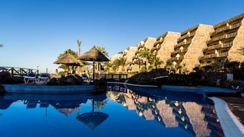 Una piscina con sillones de descanso y una sombrilla de paja, rodeada de palmeras y un edificio de varios pisos con balcones.