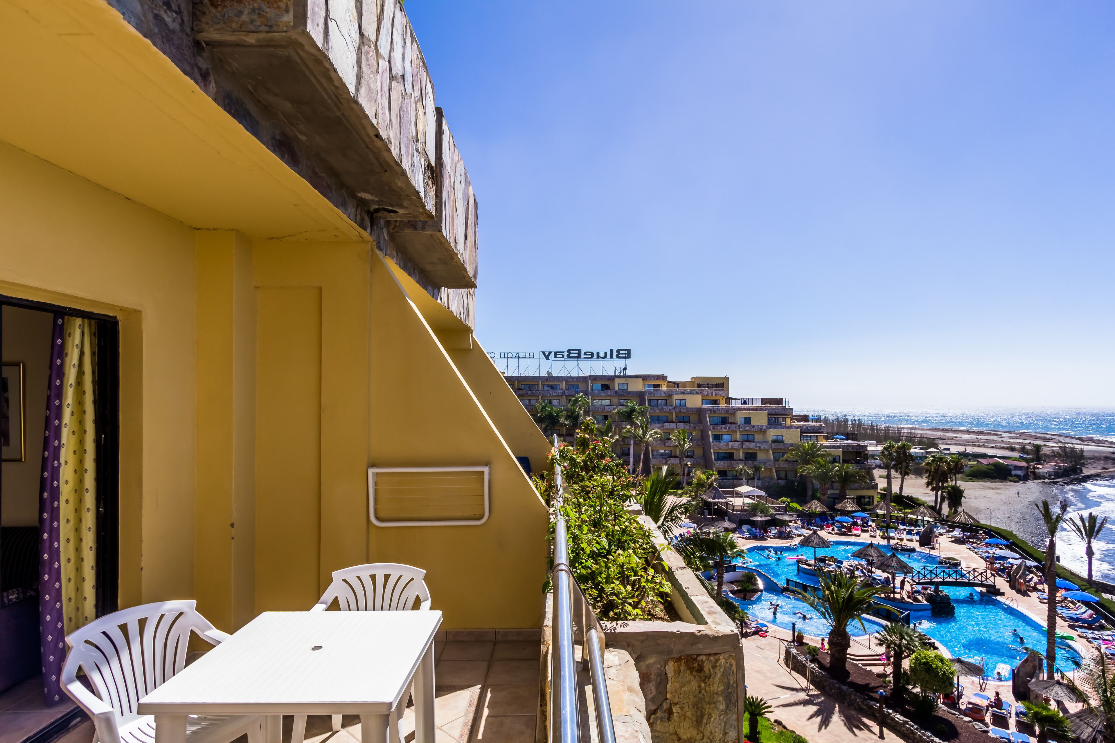 A balcony with a white table and chairs, overlooking a pool area with umbrellas and palm trees, and a view of a hotel building in the background.