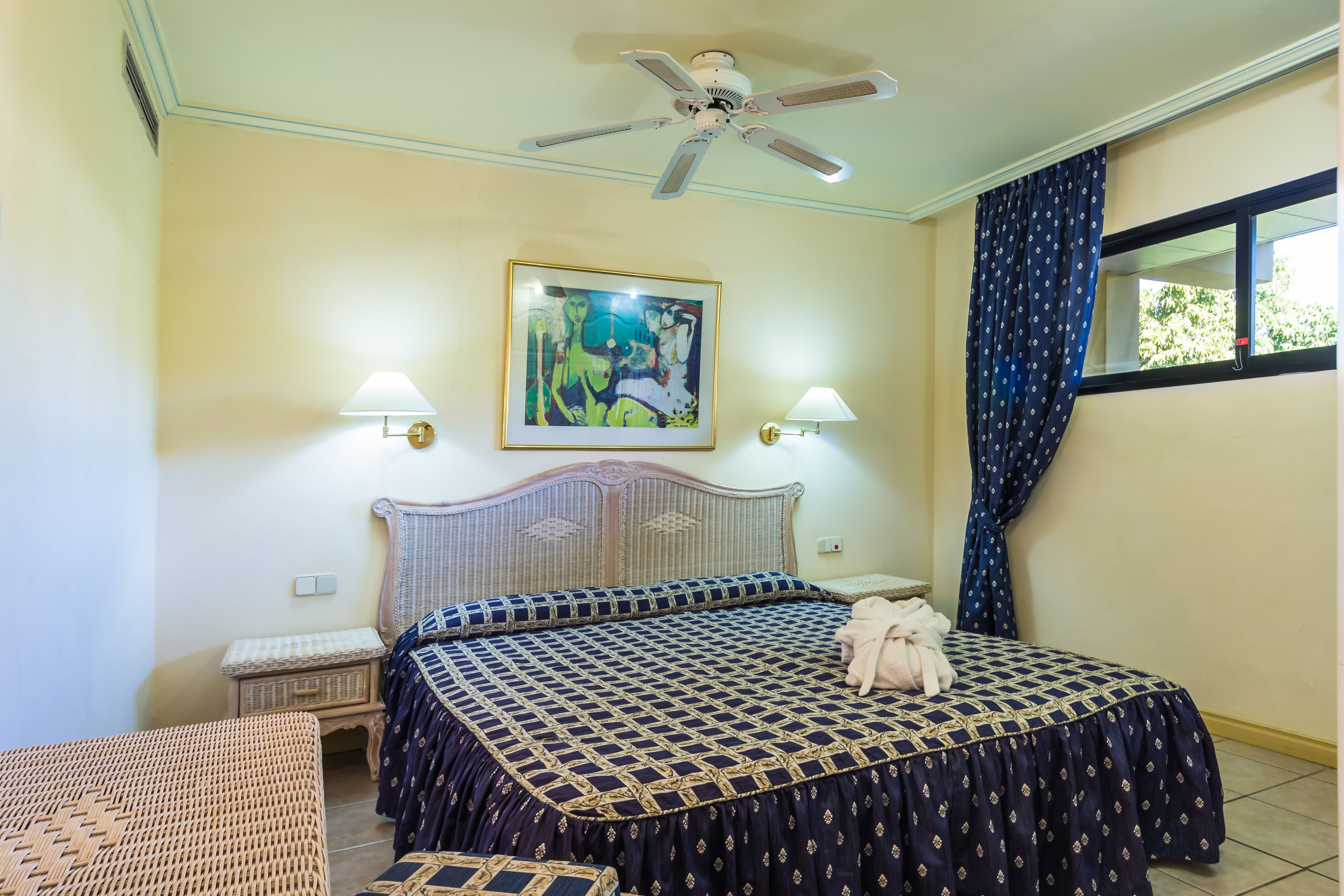 A bedroom with a bed, two bedside lamps, a ceiling fan, a window with curtains, and a painting on the wall.