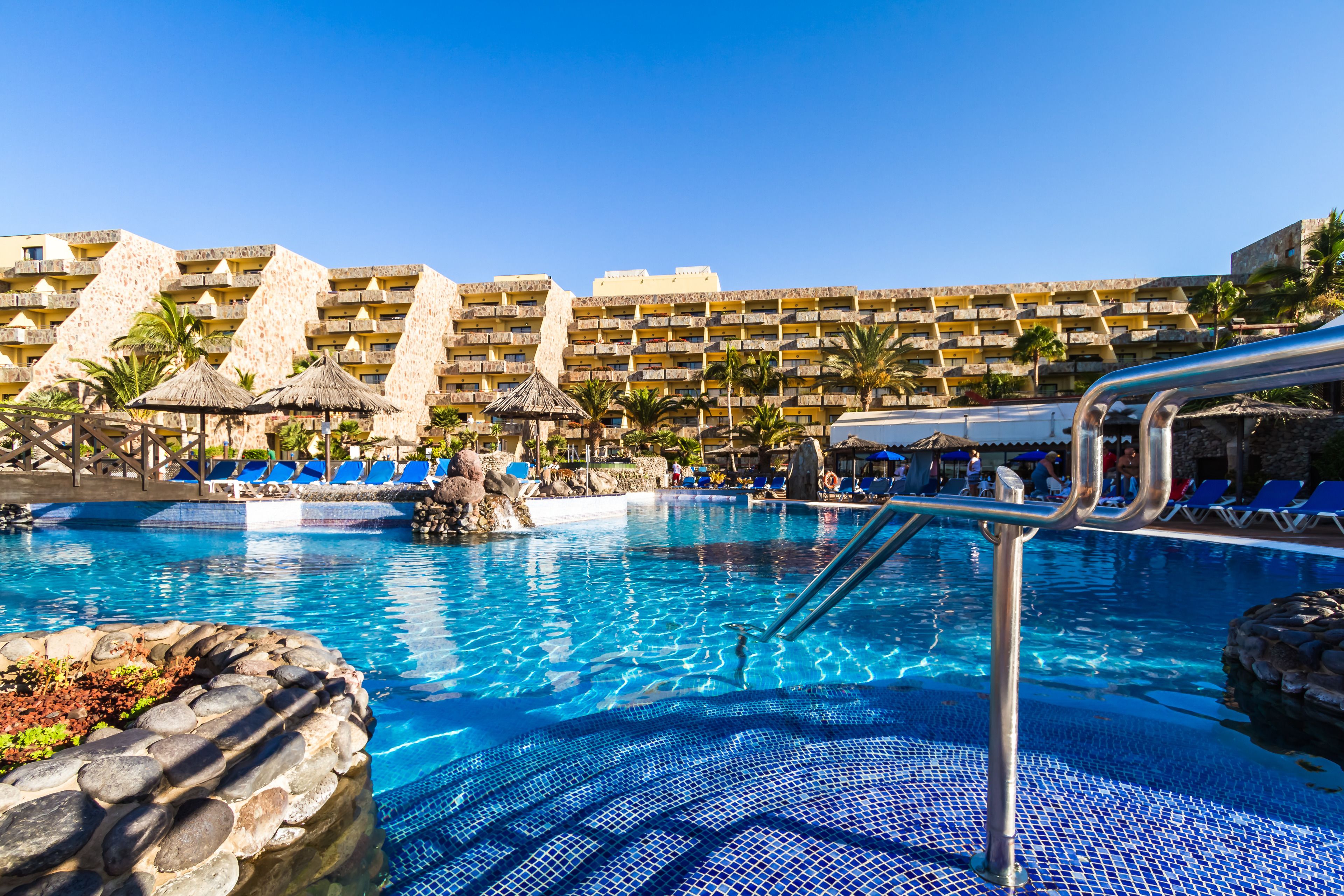A hotel pool area with clear blue water, lounge chairs, and a large building with multiple balconies in the background.