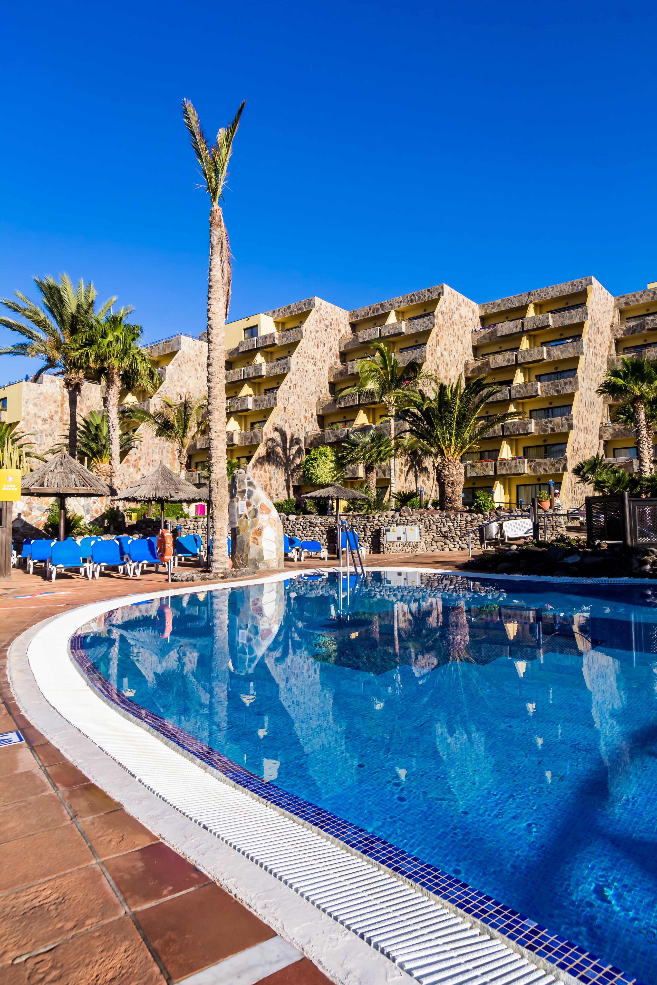 Un área de piscina con una piscina azul cristalina, sillas de descanso, palmeras y un edificio de piedra con balcones.