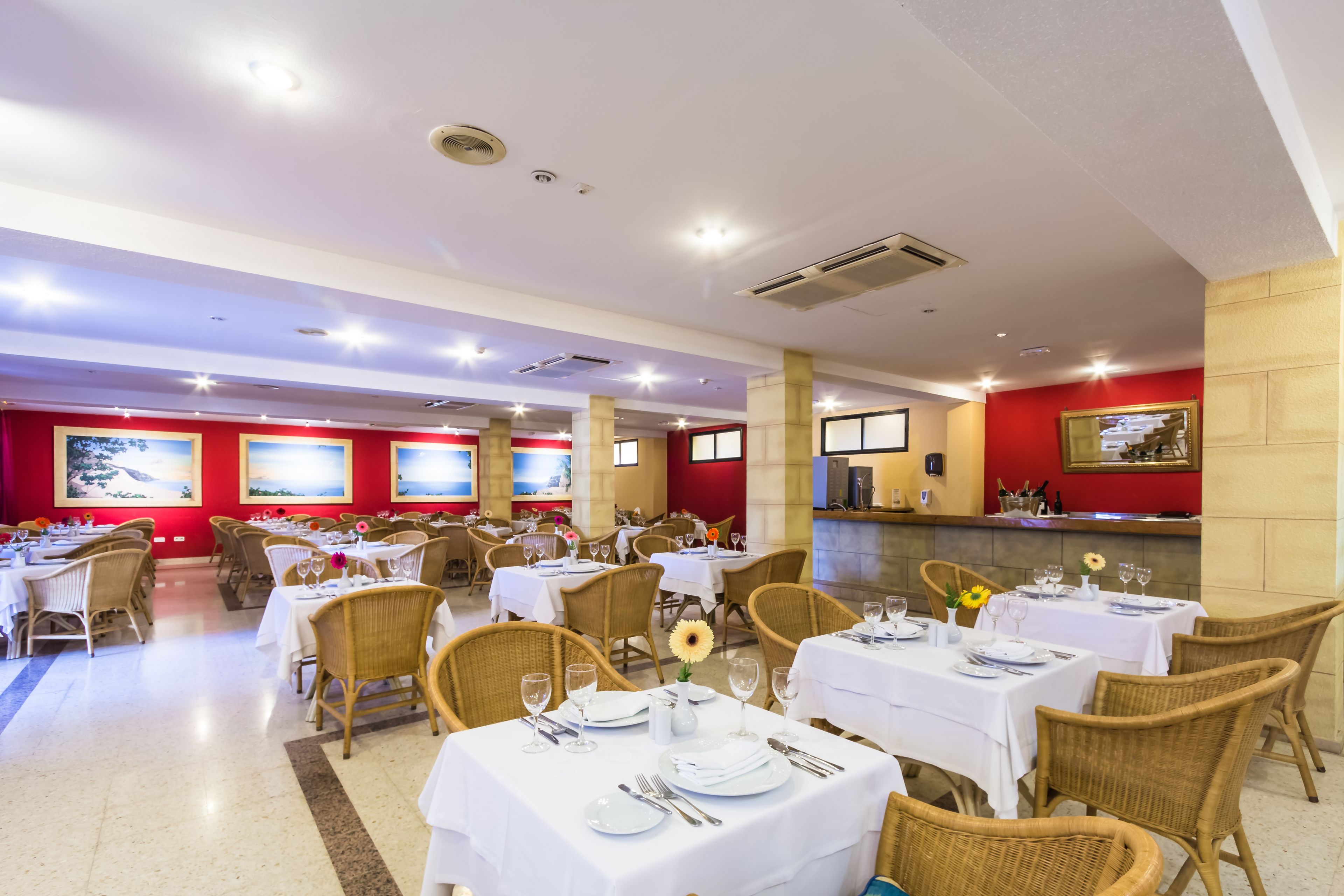 Un restaurante con varias mesas dispuestas para comer, sillas de mimbre y paredes rojas adornadas con cuadros.