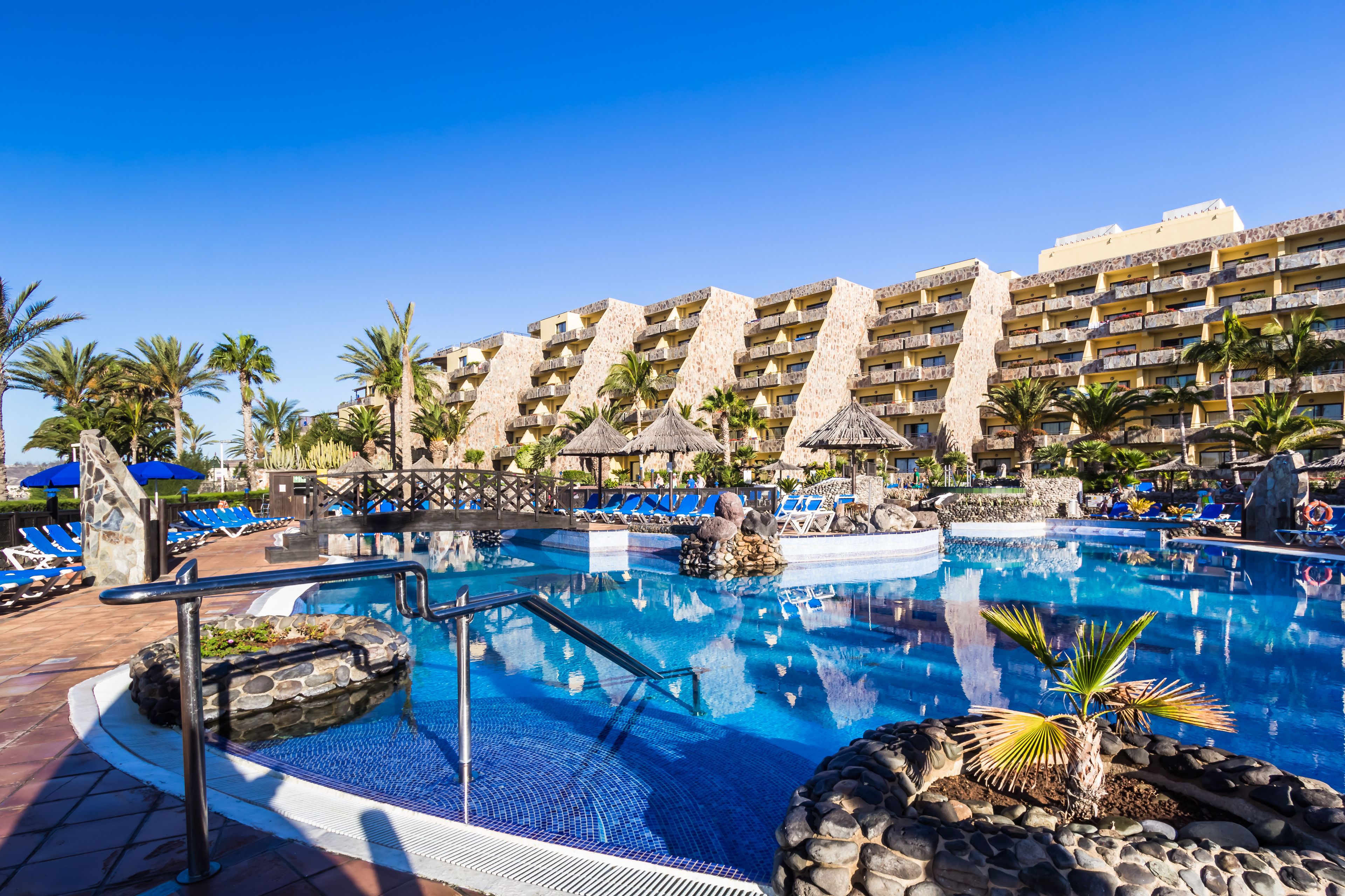 Un área de piscina de hotel con agua azul cristalina, sillas de descanso y palmeras.