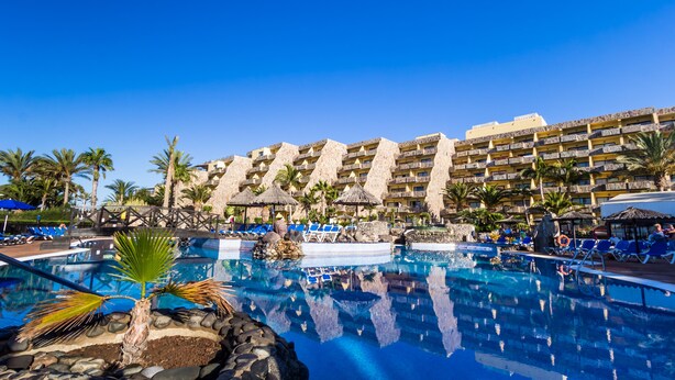 A resort pool area with clear blue water, lounge chairs, and a large building with multiple balconies. - BlueBay Beach Club Aparthotel (San Bartolomé de Tirajana)