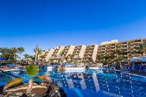 Ein Poolbereich eines Resorts mit klarem, blauem Wasser, Liegestühlen und einem großen Gebäude mit mehreren Balkonen.