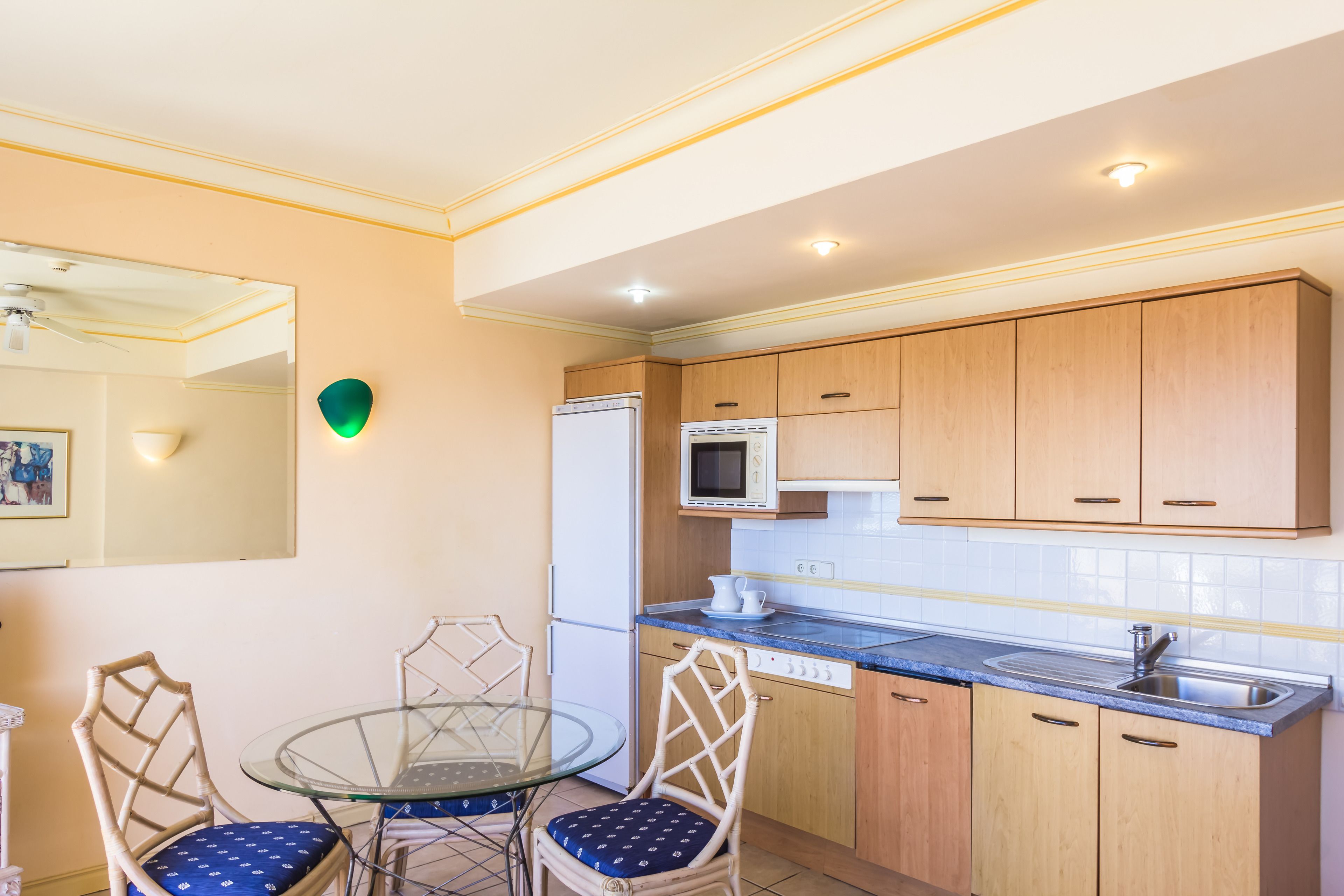 Una cocina compacta con armarios de madera, un microondas, un refrigerador y una mesa de vidrio con dos sillas.