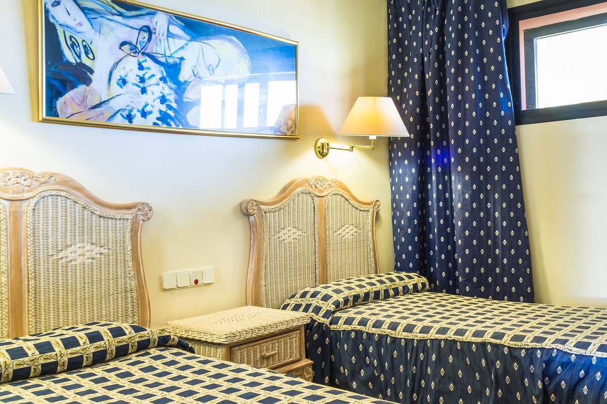 A hotel room with two beds, a ceiling fan, a painting, and a window with curtains.