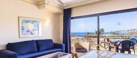A room with a sofa, a table, wicker chairs, a ceiling fan, and a large window with a view of the sea.