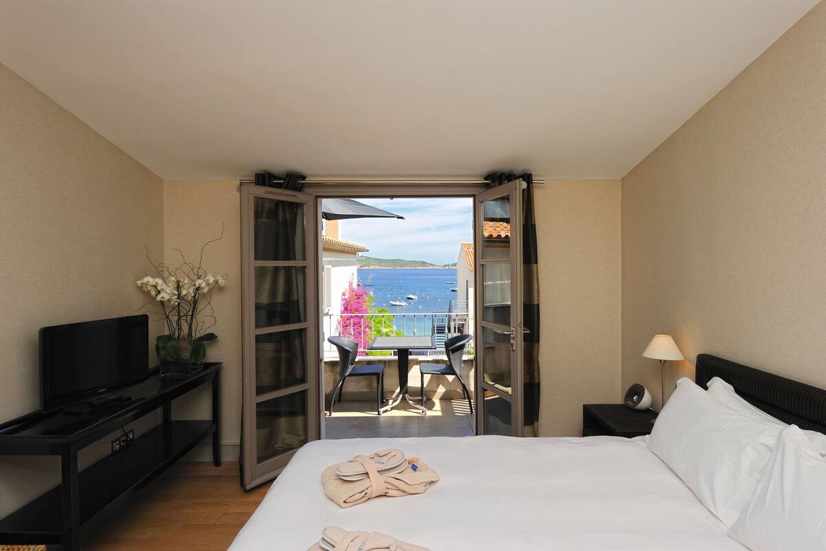 A hotel room with a bed, a TV, a desk with a chair, a balcony with a table and chairs, and a view of the sea.