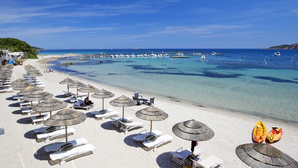 A beach with rows of sun loungers and thatched umbrellas, a clear blue sea, and boats in the distance. - Hotel Le Pinarello (Zonza)