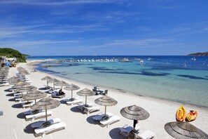 Une plage bordée de rangées de chaises longues et d’ombrelles en chaume, une mer d’un bleu limpide et des bateaux à l’horizon.