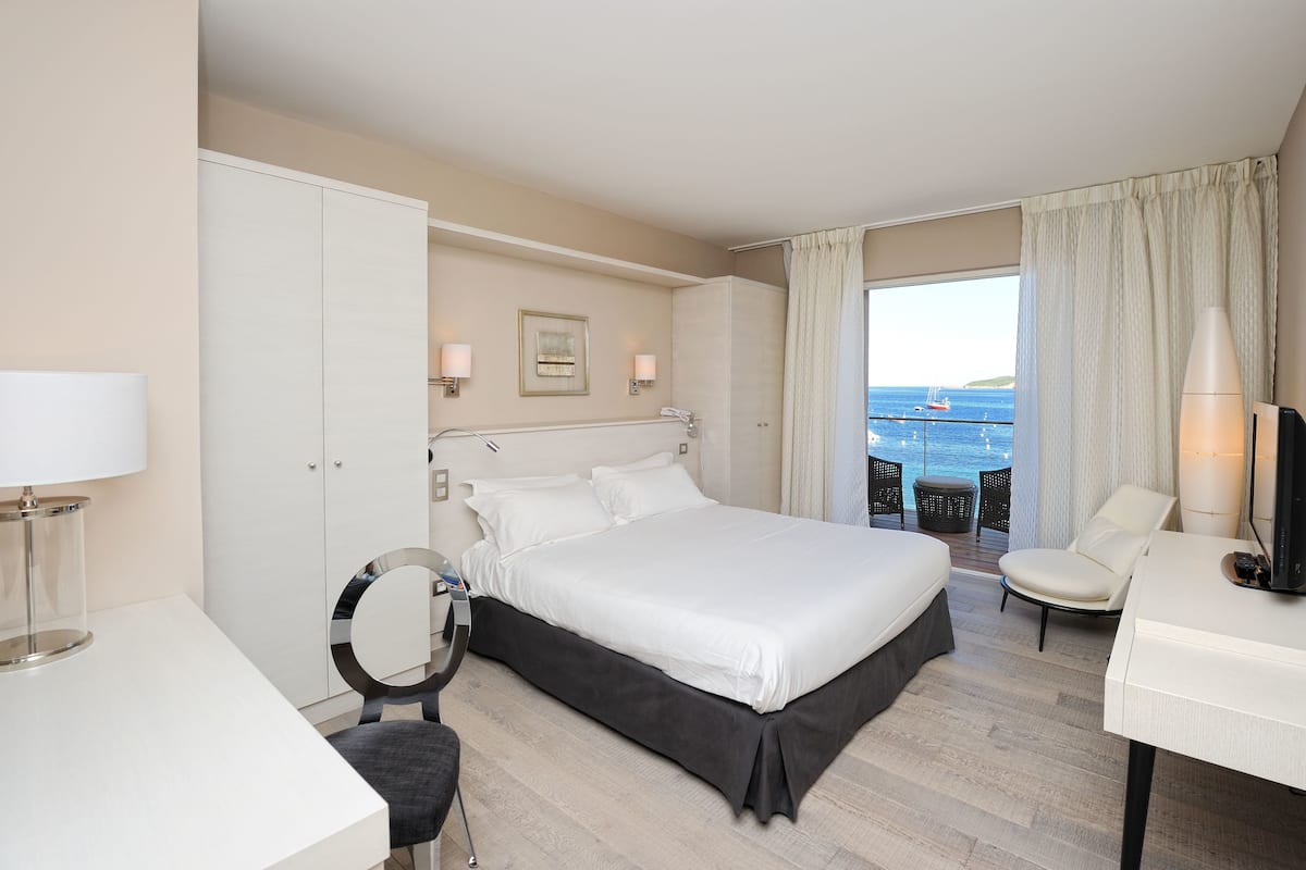 A hotel room with a large bed, a desk, a chair, a TV, a wardrobe, and a view of the sea.
