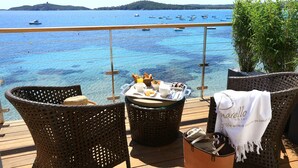 A wicker chair with a tray of breakfast items, a bucket, and a towel with hotel branding on a deck overlooking a clear blue sea with boats. - Hotel Le Pinarello (Zonza)