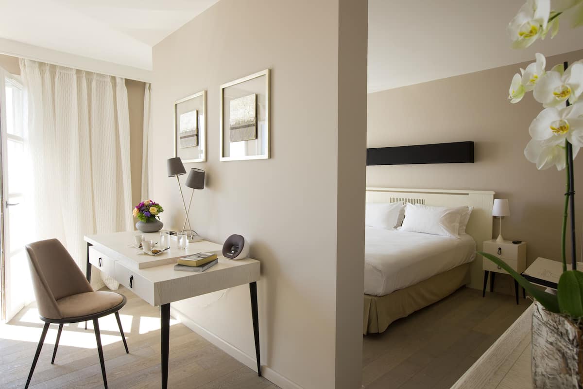 A hotel room with a bed, a desk with a chair, a nightstand, and a potted plant.