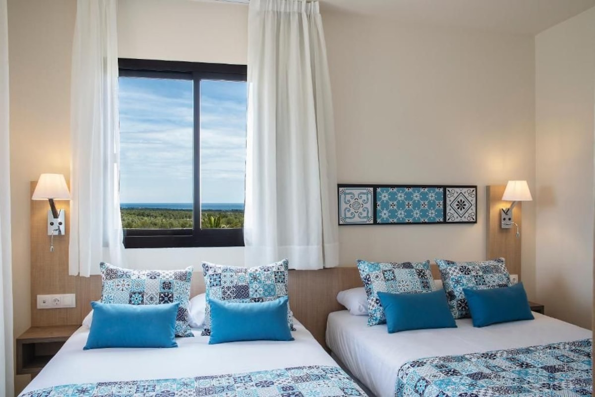 A hotel room with two beds, patterned bedding, a window with a view, and wall-mounted lamps.