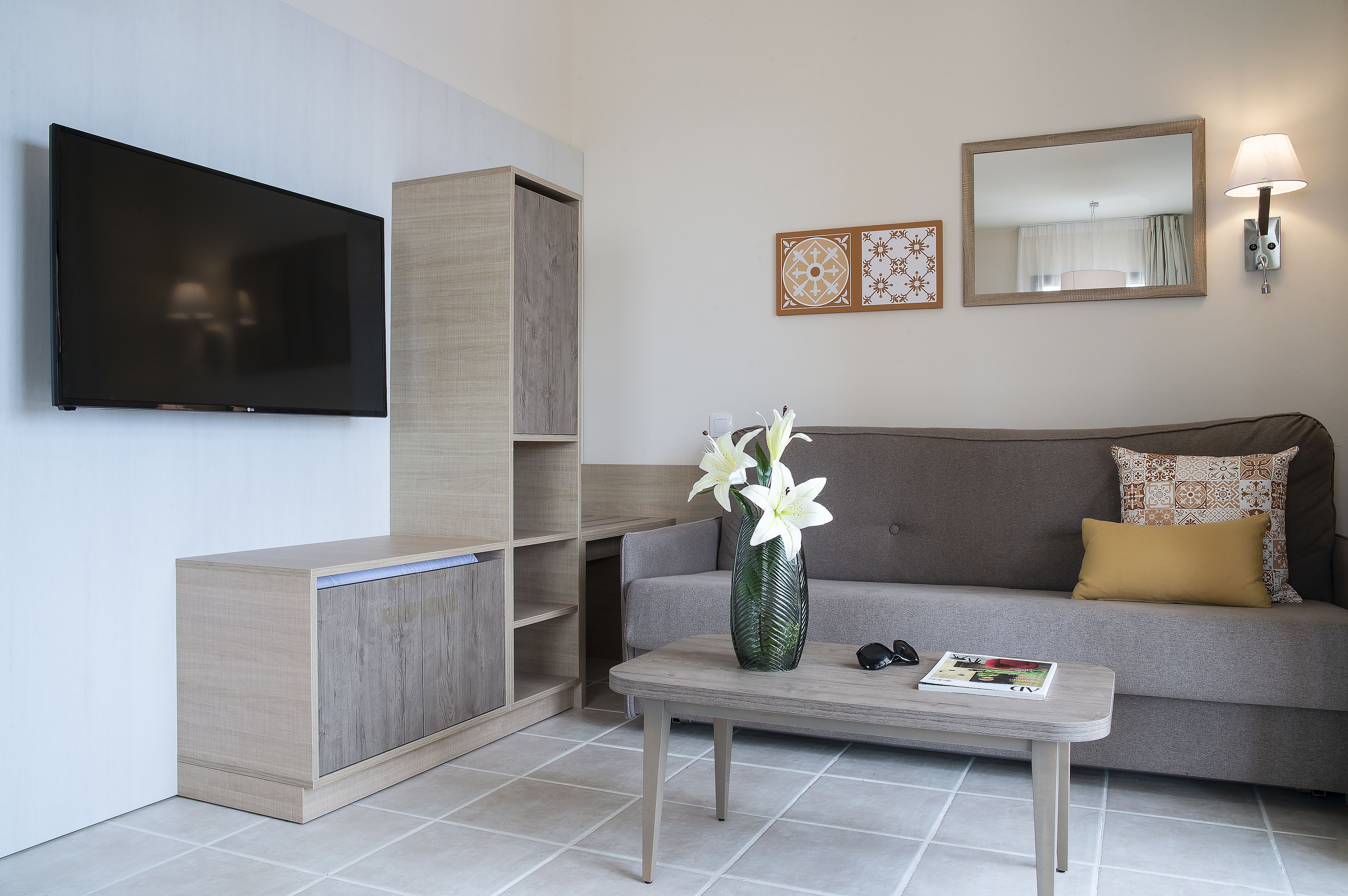 Una sala de estar moderna con una televisión de pantalla plana, un sofá con un cojín estampado, una mesa de centro con un jarrón de flores y una lámpara fijada a la pared.