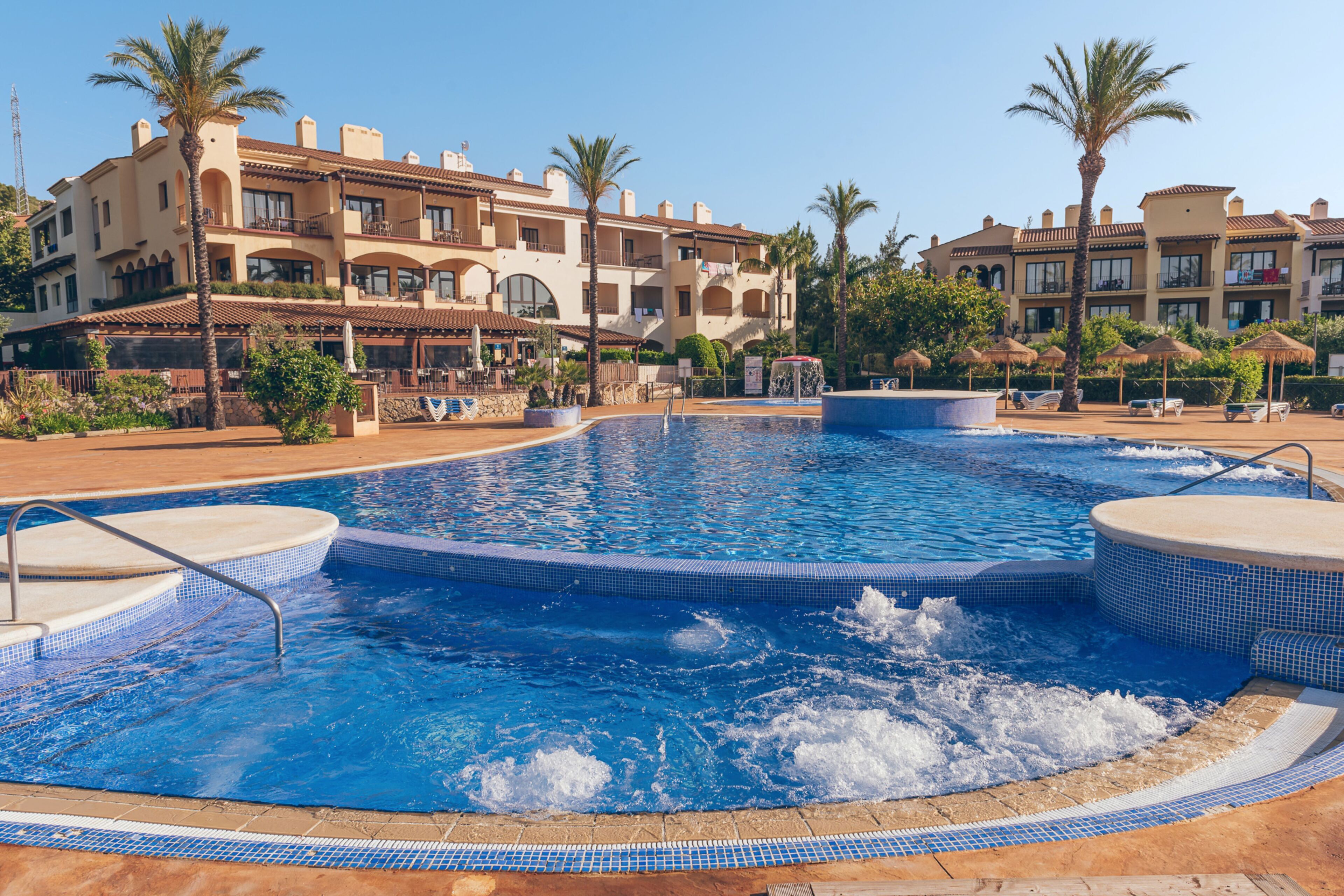 A swimming pool with a hot tub, surrounded by palm trees and a resort building with balconies.