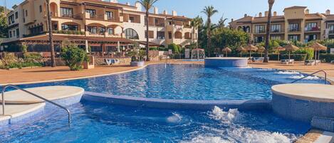A swimming pool with a hot tub, surrounded by palm trees and a resort building with balconies.