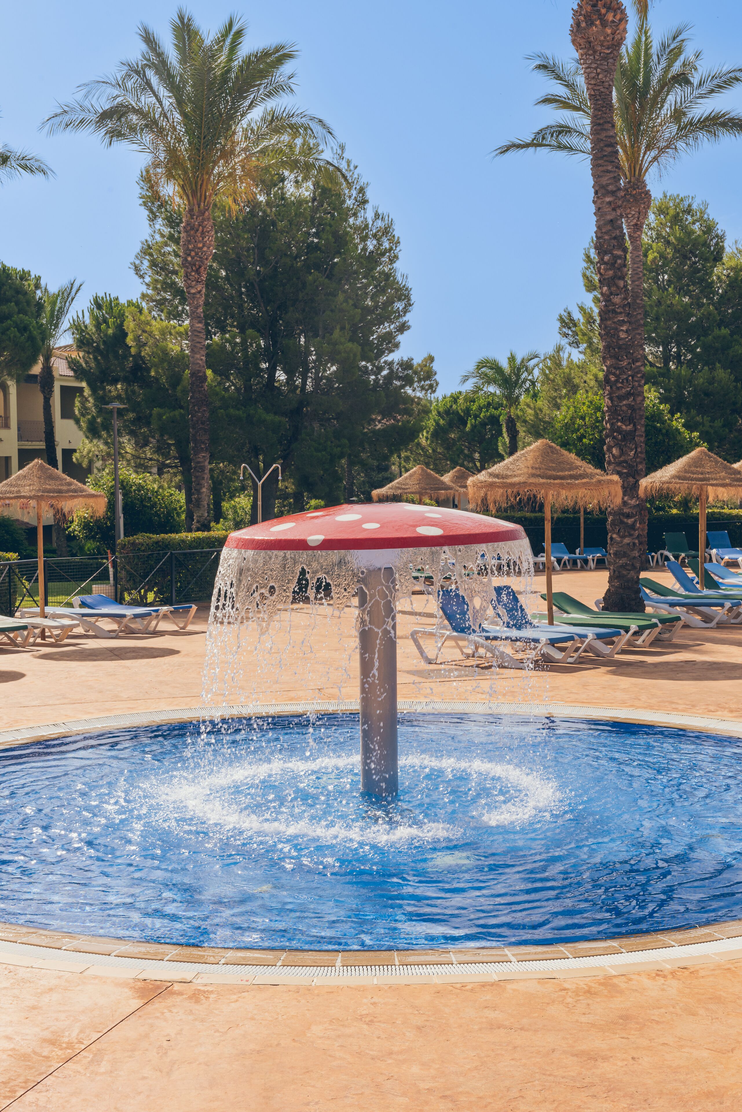 A pool with a water fountain, lounge chairs, palm trees, and thatched umbrellas.