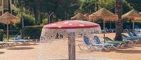 A pool with a water fountain, lounge chairs, palm trees, and thatched umbrellas.