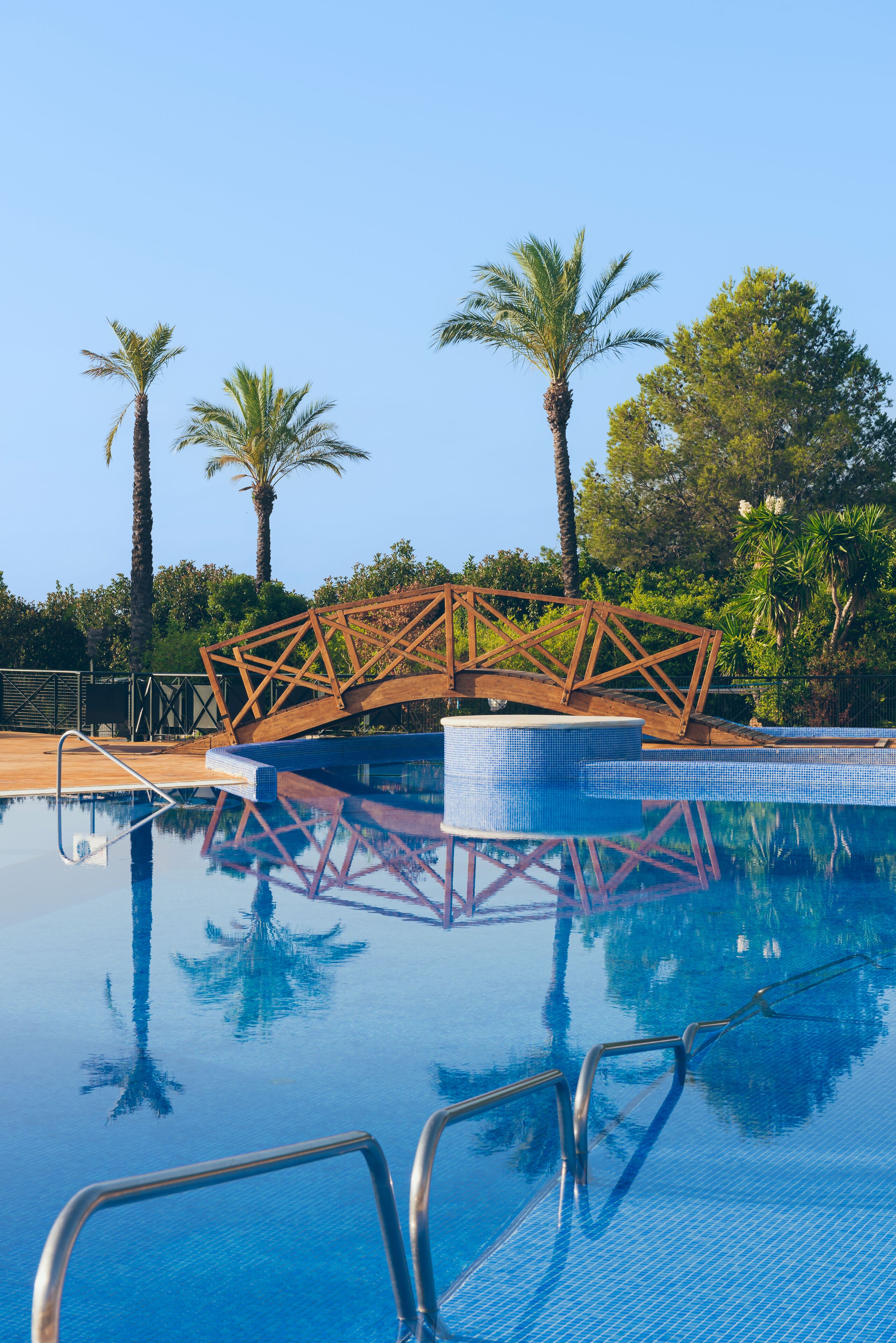 Una piscina con un puente de madera, rodeada de palmeras y cielos despejados.