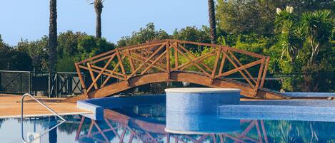 Una piscina con un puente de madera, rodeada de palmeras y cielos despejados.