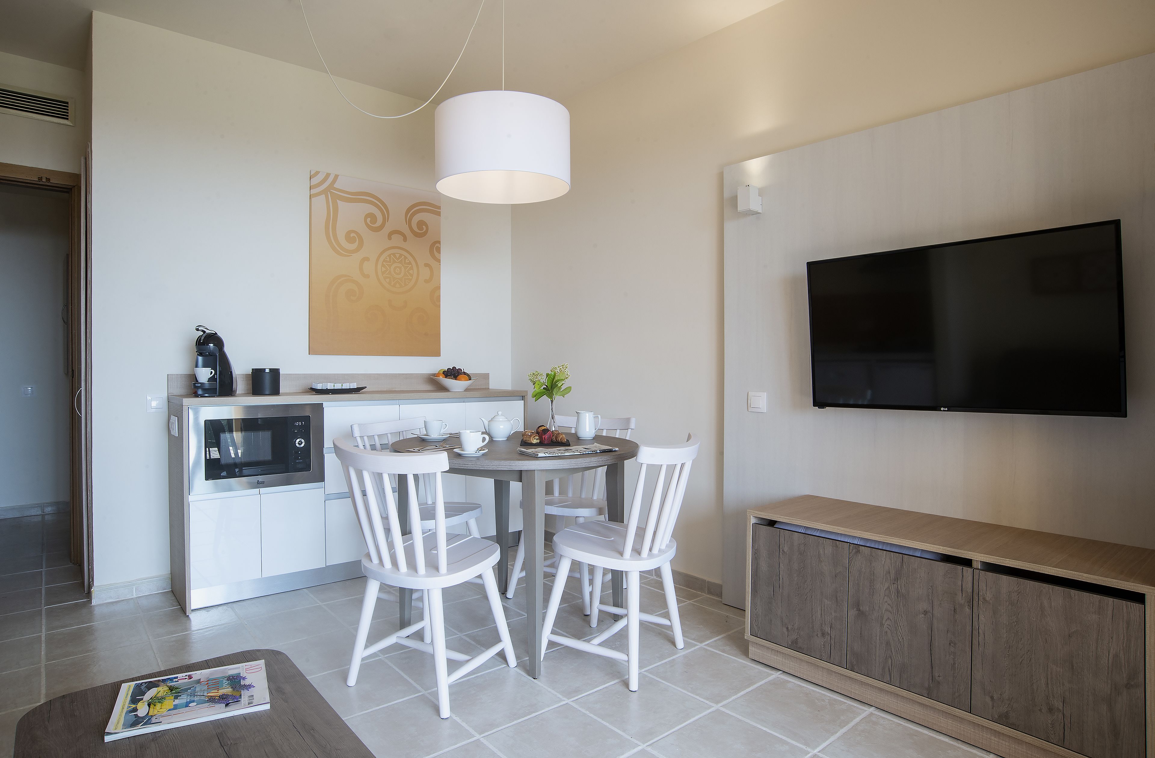 Una cocina moderna con microondas, hervidor y una mesa de comedor con sillas. Un televisor de pantalla plana está montado en la pared sobre la encimera.