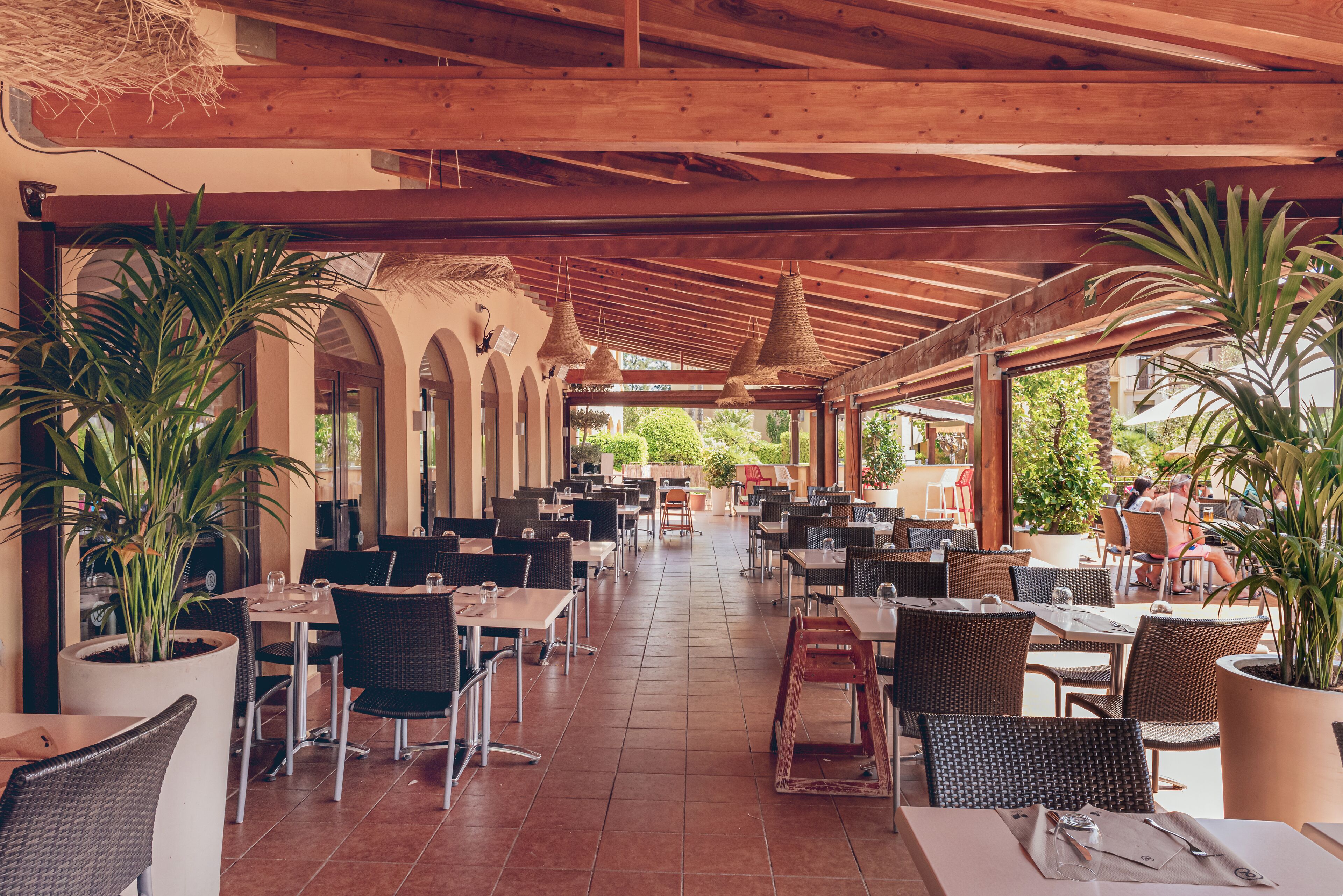 An outdoor cafe with multiple tables and chairs, potted plants, and a wooden ceiling structure.