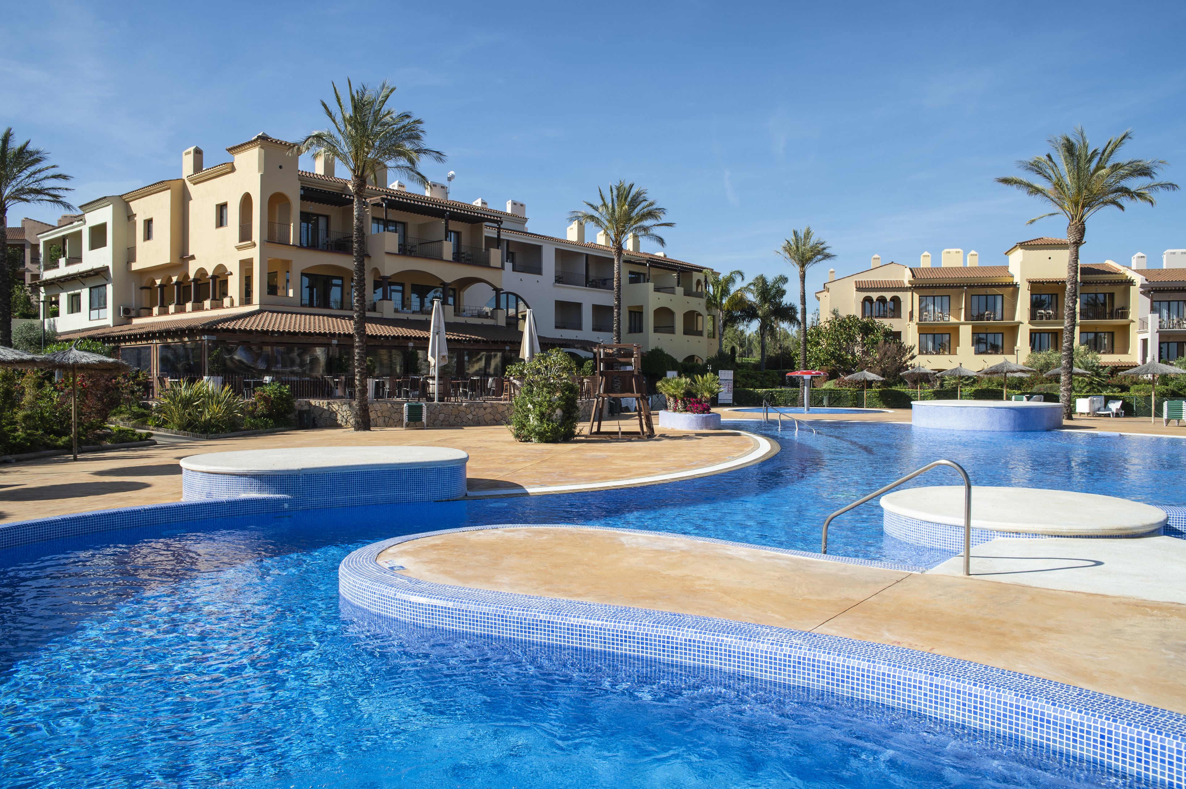 Een hotel met een zwembad, palmbomen en een mediterrane architectuur.